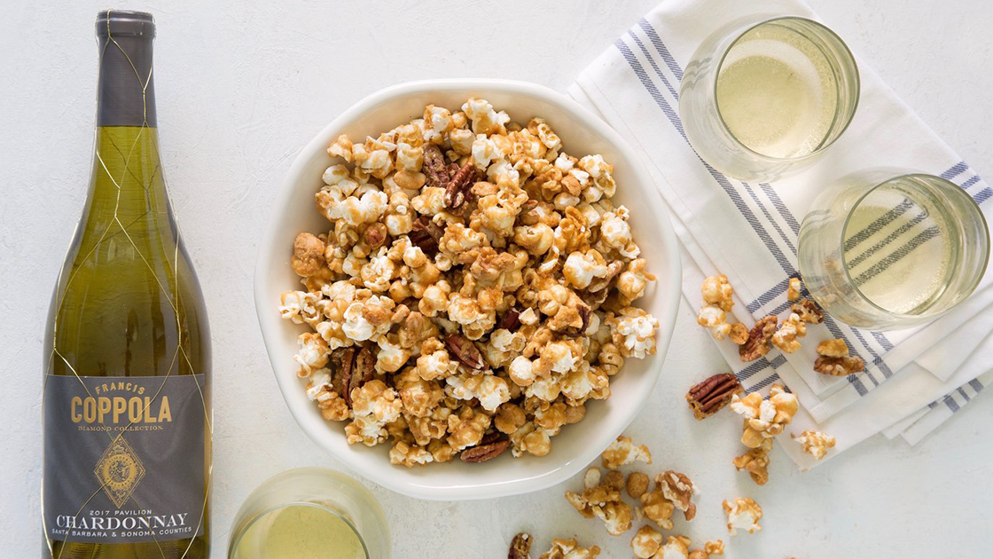 Large bowl of caramel corn with a bottle of chardonnay and three glasses.