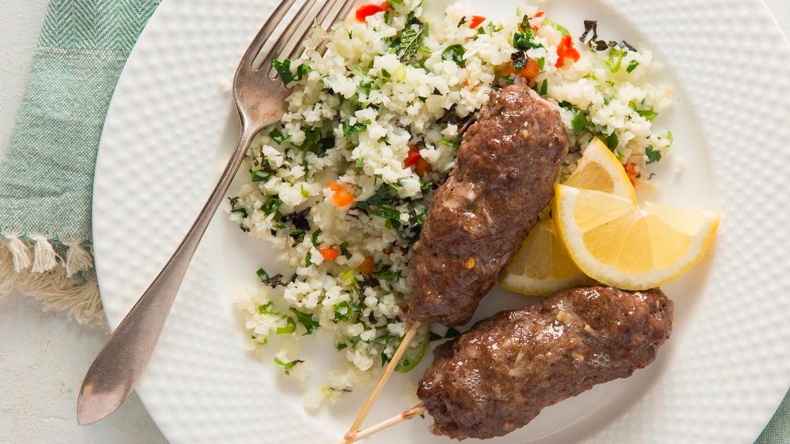 Two Greek beef skewers on a plate with lemon wedges, a fork and cauliflower tabbouleh.