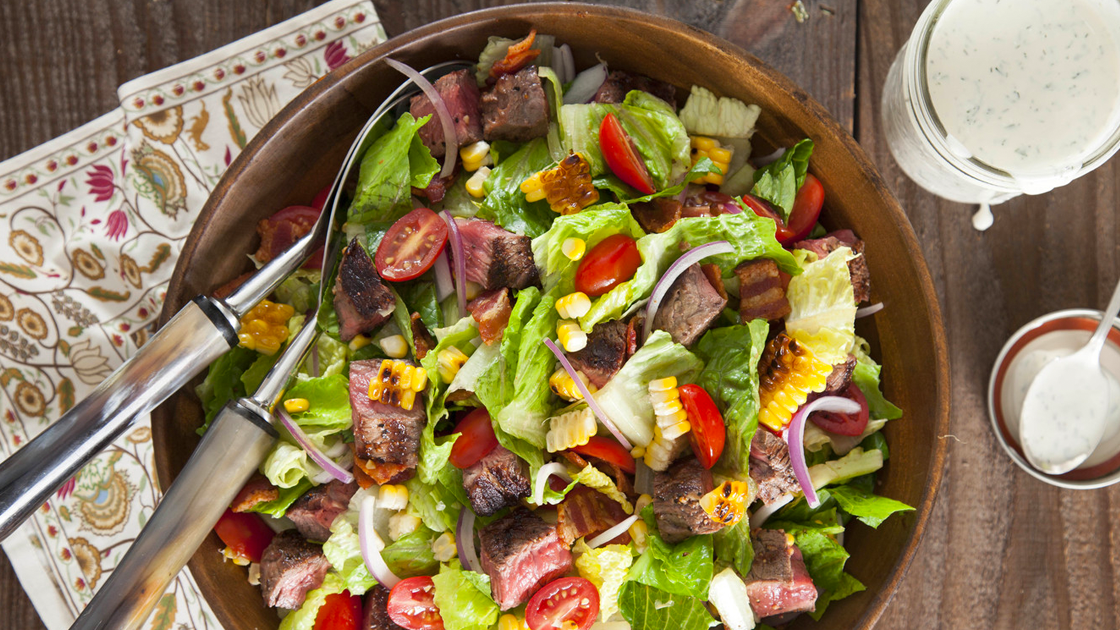 A large salad topped with cubed steak, tomatoes, corn and onion in a wooden bowl next to jars of ranch dressing.