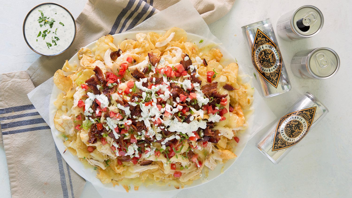 Plate full of nachos made with kettle chips, a bowl of ranch dressing and cans of Diamond wines.