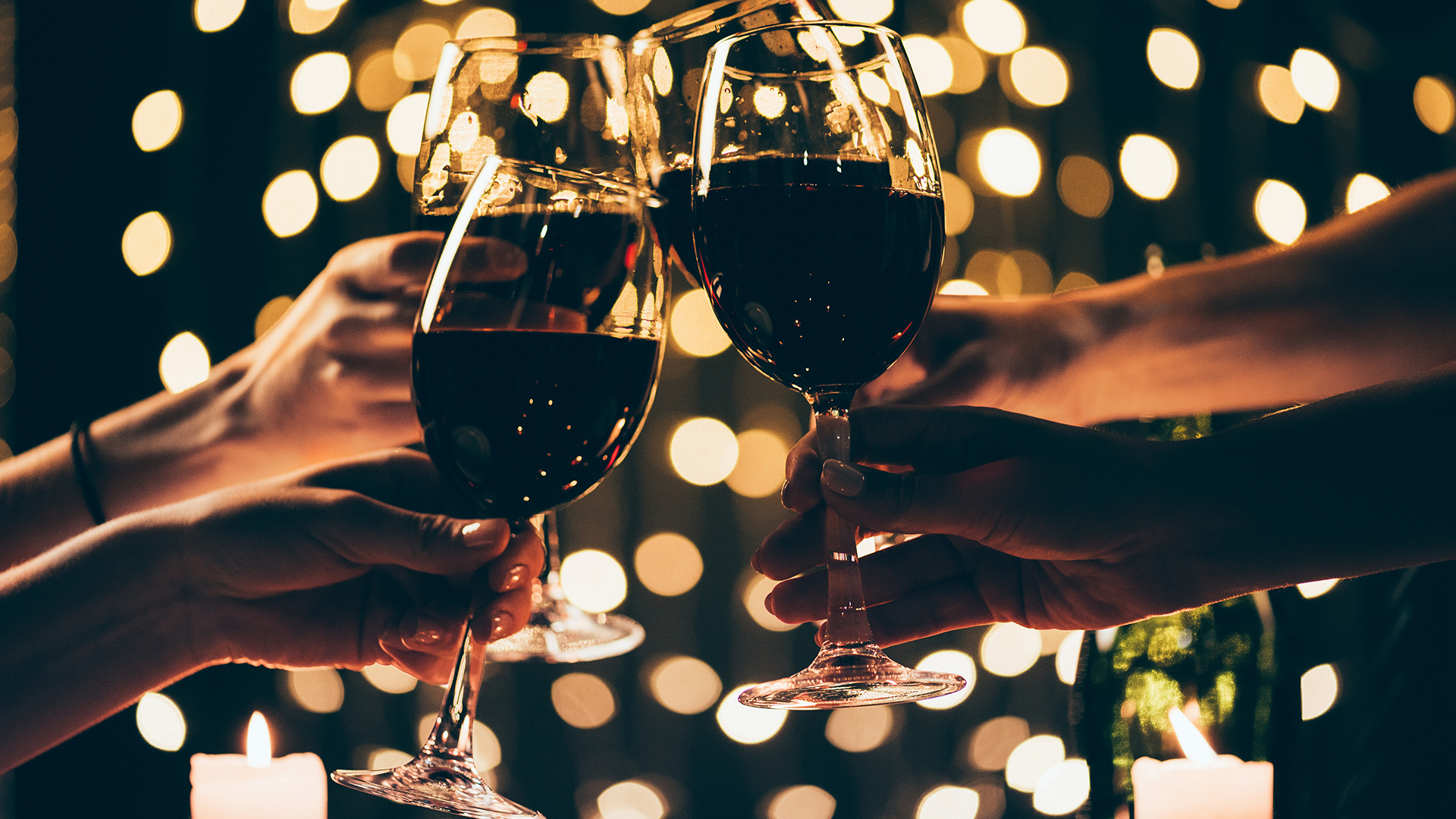 Four hands toasting with glasses of red wine with string lights in the background.