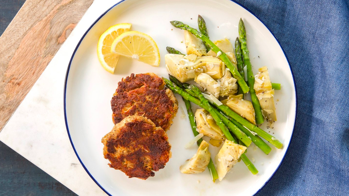 A plate with tune cakes, lemon wedges and a artichoke hearts and asparagus melody.